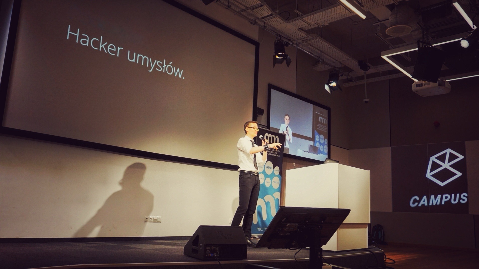 Ludwik C. Siadlak w białej koszuli i krawacie przemawia na scenie Google Campus Warsaw, na ekranie napis „Hacker umysłów”